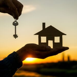Hands holding a key and a model home in front of a sunset